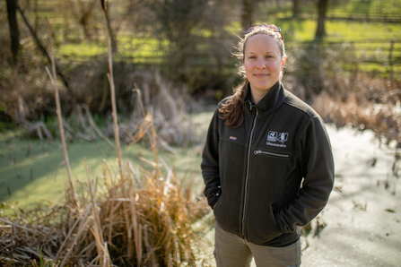Nicola looking at the camera standing in front of a pond, wearing a GWT fleece