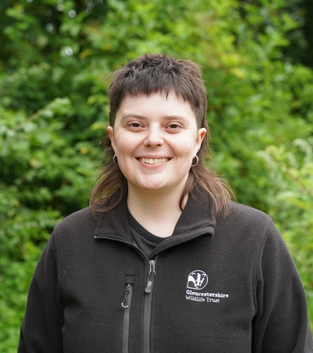 A photo of Sophie, Gloucestershire Wildlife Trust's Head of External Relations. She is looking straight at the camera and smiling, wearing a GWT fleece with a backdrop of greenery. 